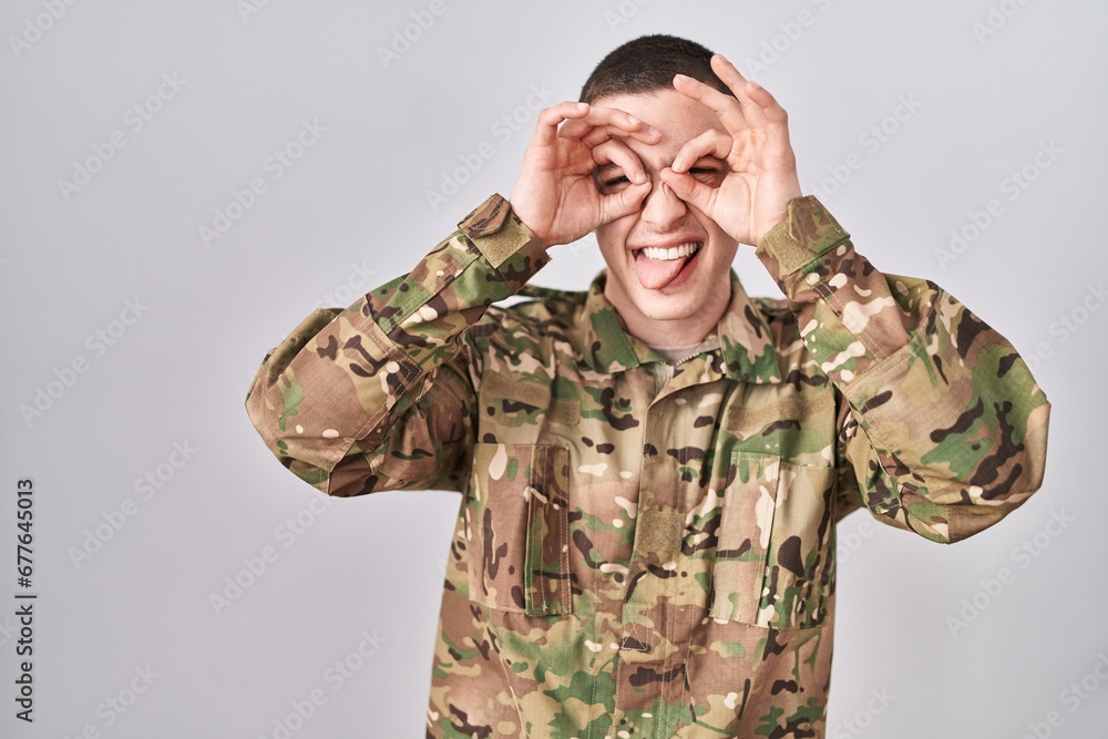 Young man wearing camouflage army uniform doing ok gesture like binoculars sticking tongue out, eyes looking through fingers. crazy expression.