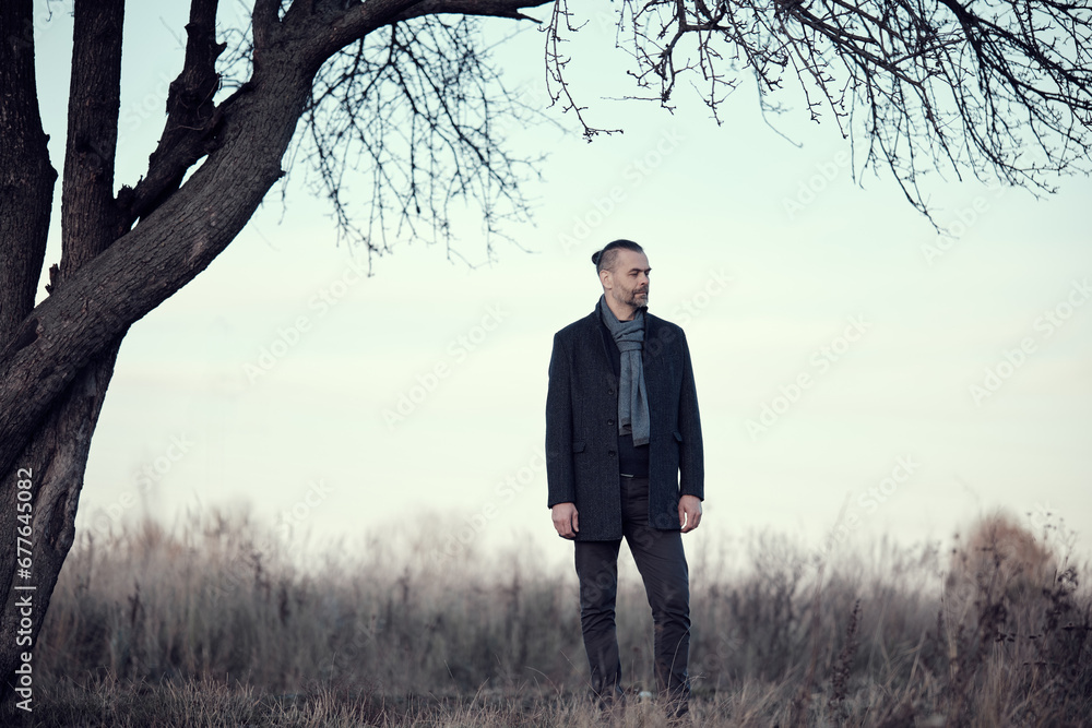 Portrait of a bearded middle-aged man in a blue coat and wool scarf standing amidst a natural landscape near a tree in the autumn and winter season. Portrait of a bearded middle-aged man in a blue coat and wool scarf standing amidst a natural landscape near a tree in the autumn and winter season.
