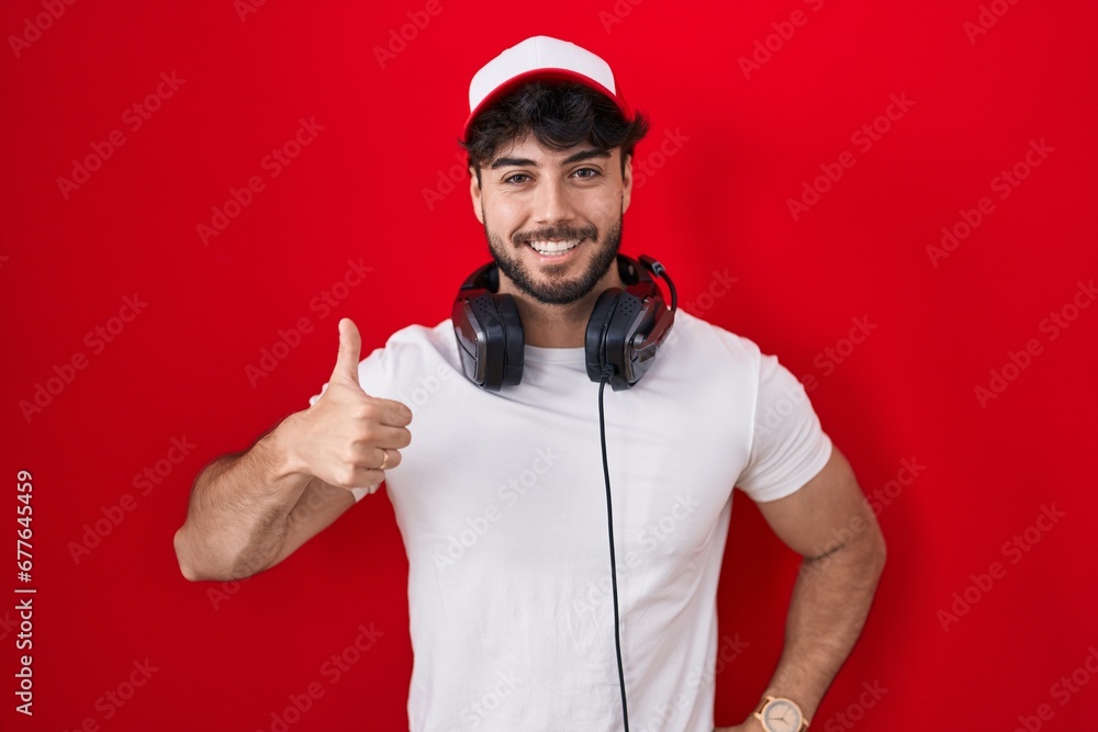 Hispanic man with beard wearing gamer hat and headphones doing happy thumbs up gesture with hand. approving expression looking at the camera showing success. Hispanic man with beard wearing gamer hat and headphones doing happy thumbs up gesture with hand. approving expression looking at the camera showing success.