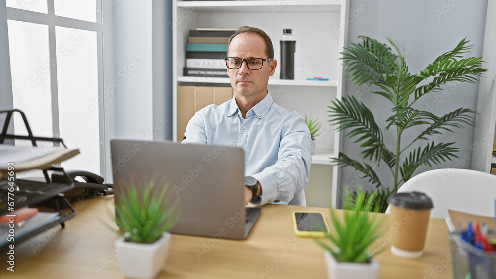 In the heart of his workplace, a mature hispanic man with a serious face, works hard on his laptop in the office In the heart of his workplace, a mature hispanic man with a serious face, works hard on his laptop in the office