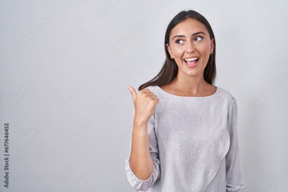 Young hispanic woman standing over white background smiling with happy face looking and pointing to the side with thumb up. Young hispanic woman standing over white background smiling with happy face looking and pointing to the side with thumb up.