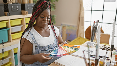 Serious african american woman artist, with beautiful black braids, choosing vibrant color palette at art studio amidst painting lesson