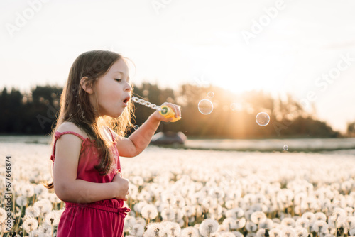 Playful lady standing in white dandelions meadow and blowing soap bubbles at sunset. Child enjoying summer in nature.