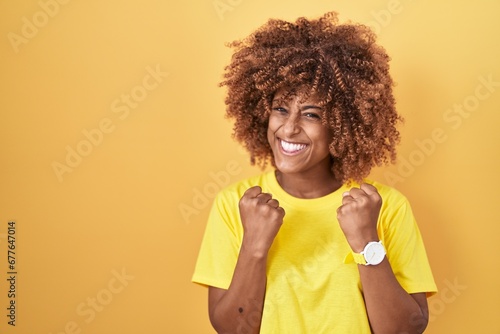 Young hispanic woman with curly hair standing over yellow background very happy and excited doing winner gesture with arms raised, smiling and screaming for success. celebration concept.