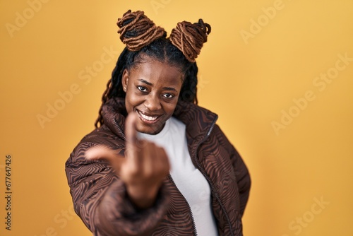 African woman with braided hair standing over yellow background beckoning come here gesture with hand inviting welcoming happy and smiling photo