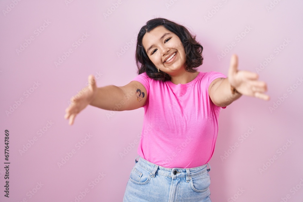 Young hispanic woman standing over pink background looking at the camera smiling with open arms for hug. cheerful expression embracing happiness. Young hispanic woman standing over pink background looking at the camera smiling with open arms for hug. cheerful expression embracing happiness.