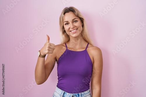 Young blonde woman standing over pink background doing happy thumbs up gesture with hand. approving expression looking at the camera showing success. © Krakenimages.com