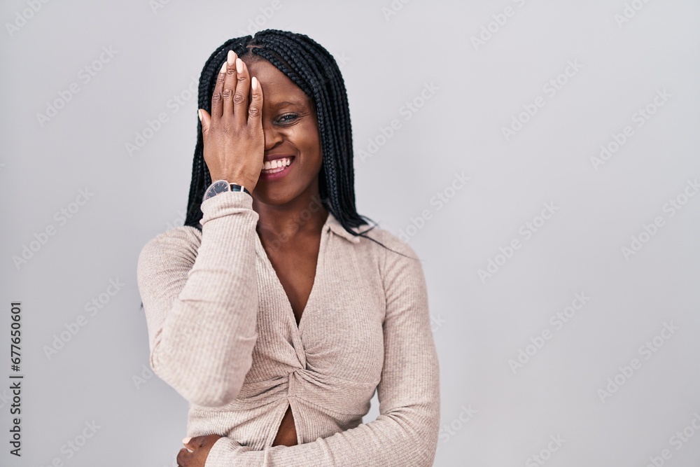 African woman with braids standing over white background covering one eye with hand, confident smile on face and surprise emotion. African woman with braids standing over white background covering one eye with hand, confident smile on face and surprise emotion.