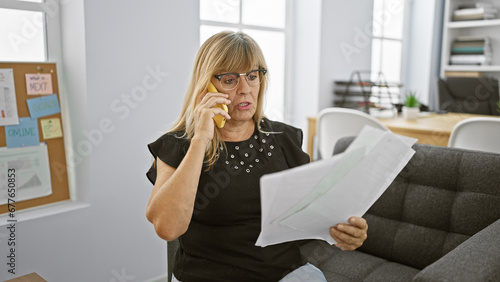 Elegant blonde boss woman in her middle age - relaxed, yet seriously engrossed in a business conversation on her smartphone, while diligently reading an important office document.