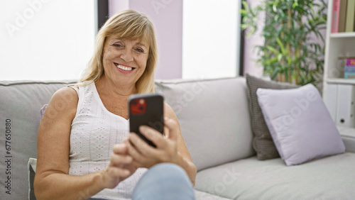 Beautiful middle-aged blonde woman sitting on a sofa at home, enjoying the joy of texting on her smartphone, a cheerful expression of happiness on her face