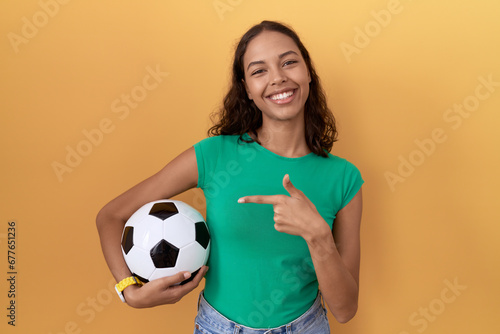 Young hispanic woman holding ball cheerful with a smile on face pointing with hand and finger up to the side with happy and natural expression © Krakenimages.com