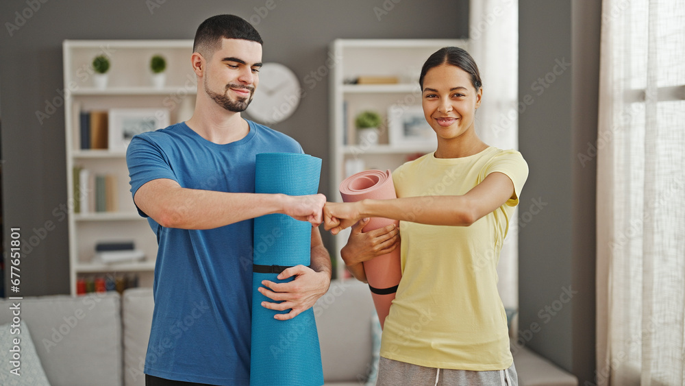 Obraz premium Beautiful couple bumping fists, holding a yoga mat, smiling with fitness glee in the home living room