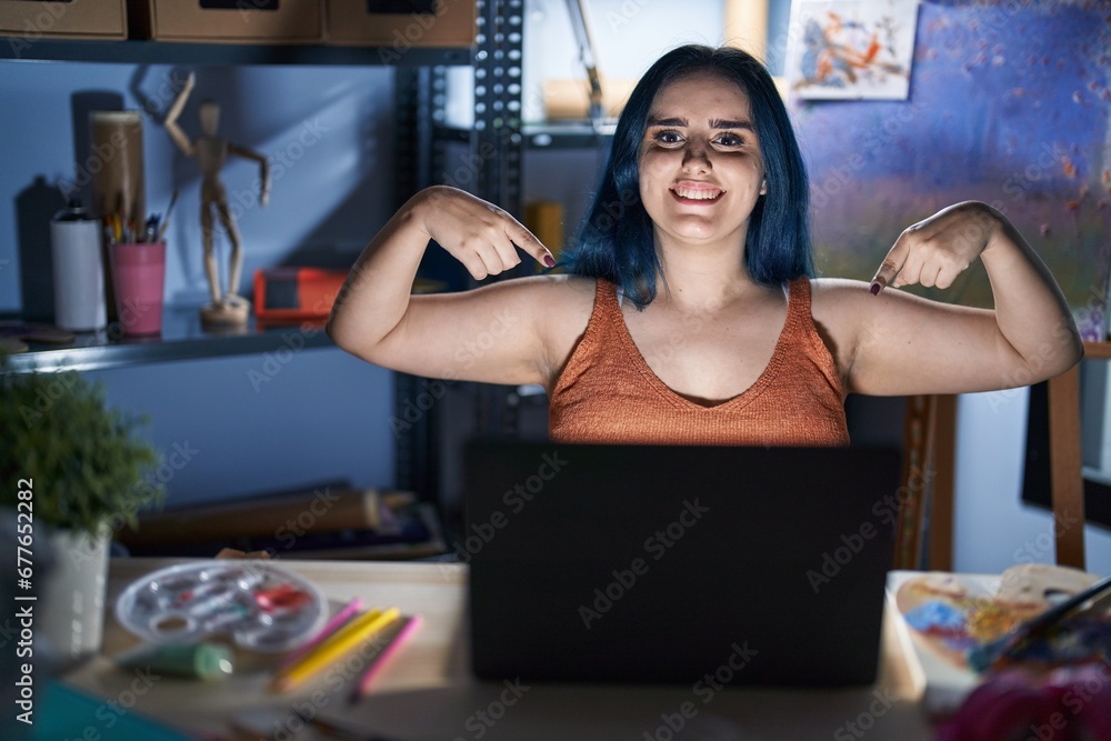 Young modern girl with blue hair sitting at art studio with laptop at night looking confident with smile on face, pointing oneself with fingers proud and happy. Young modern girl with blue hair sitting at art studio with laptop at night looking confident with smile on face, pointing oneself with fingers proud and happy.