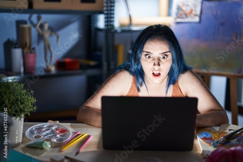 Young modern girl with blue hair sitting at art studio with laptop at night afraid and shocked with surprise expression, fear and excited face.