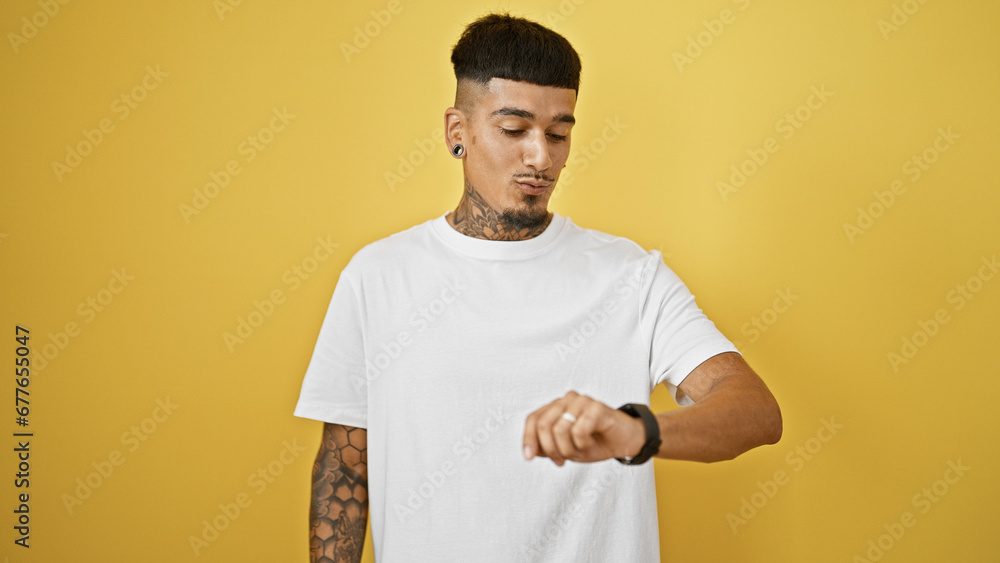 Handsome young latin man, tattooed and relaxed, looking at his wristwatch with a serious expression. he's standing against a cool, isolated yellow background, countdown timer in focus. Handsome young latin man, tattooed and relaxed, looking at his wristwatch with a serious expression. he's standing against a cool, isolated yellow background, countdown timer in focus.
