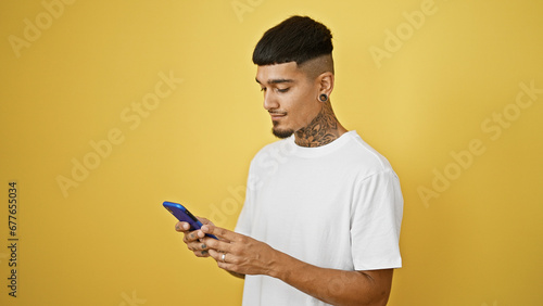 Cool tattooed young latin man concentrates on texting, his serious expression illuminated by isolated yellow background © Krakenimages.com