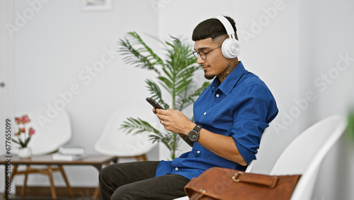 Young latin man, engrossed in his phone, tapping to the rhythm echoing through his headphones, ensconced on chair in the waiting room