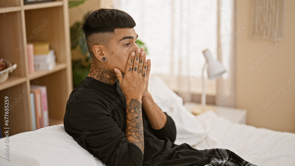 Stressed young latin man sitting on bed, wrestling with anxiety as morning breaks in his bedroom, mirroring the emotional struggle within his tattooed portrait, manifesting as a throbbing headache