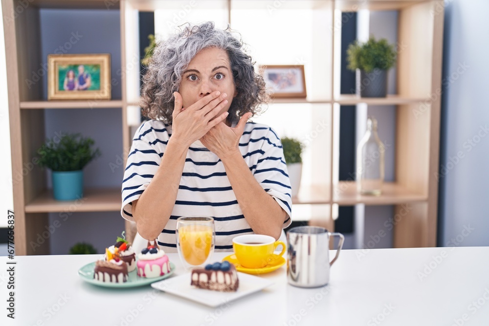 Middle age woman with grey hair eating pastries and drinking coffee for breakfast shocked covering mouth with hands for mistake. secret concept. Middle age woman with grey hair eating pastries and drinking coffee for breakfast shocked covering mouth with hands for mistake. secret concept.