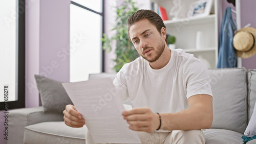 Young hispanic man reading document sitting on sofa at home