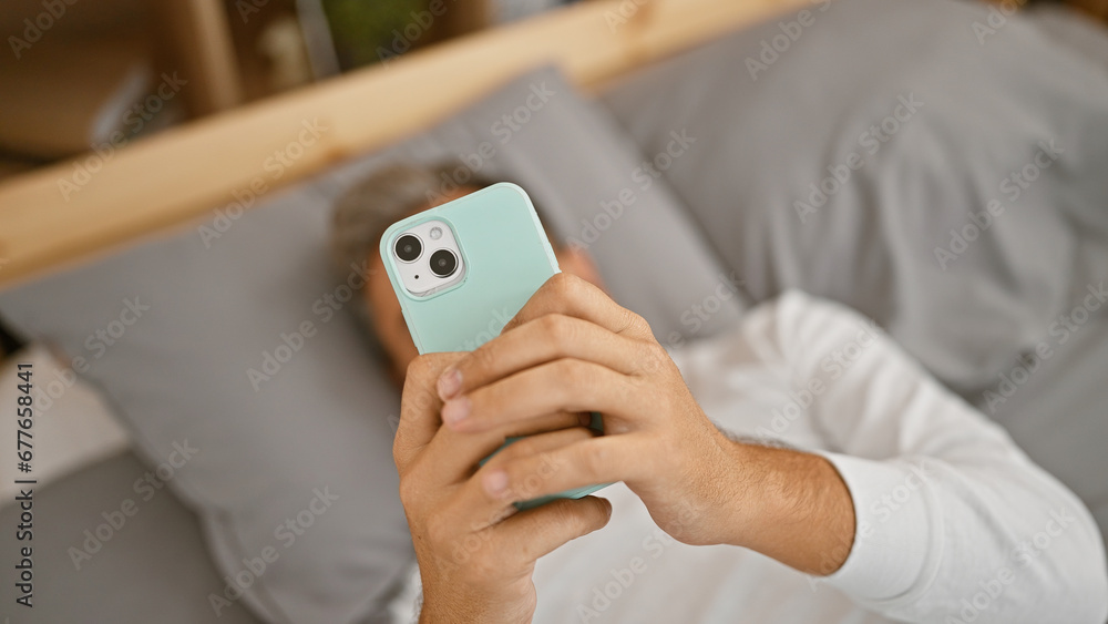 Young, handsome hispanic man with grey hair, comfortably chilling in bed, engrossed in texting on his smartphone. a relaxed morning ambiance fills his cozy bedroom. Young, handsome hispanic man with grey hair, comfortably chilling in bed, engrossed in texting on his smartphone. a relaxed morning ambiance fills his cozy bedroom.