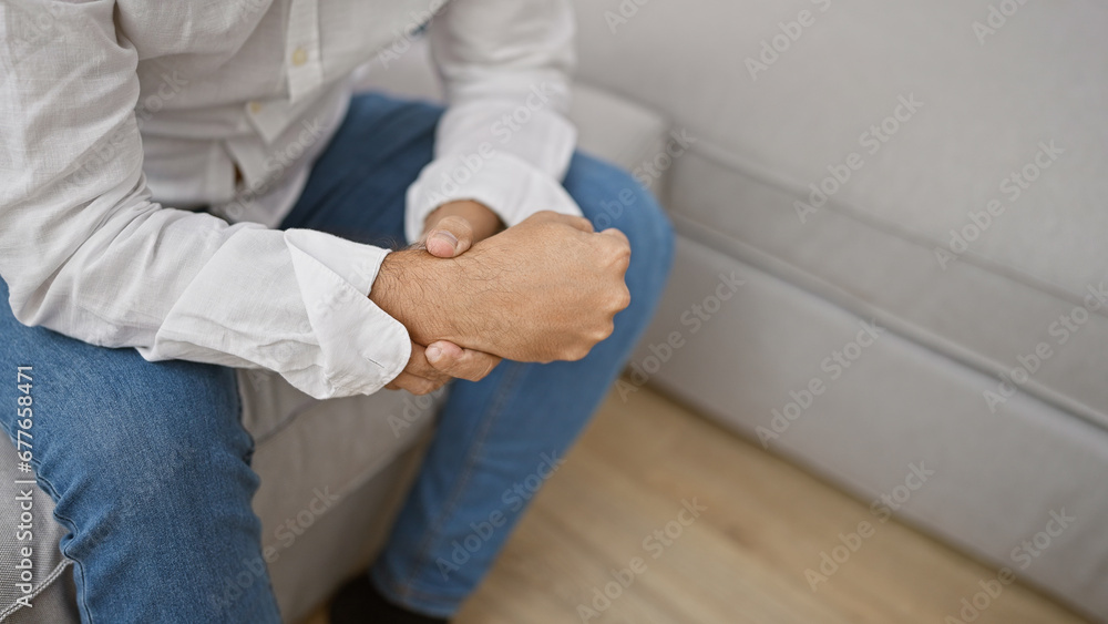 Worried young hispanic man with grey hair, enduring wrist pain at home, sitting unhappily on his living room sofa Worried young hispanic man with grey hair, enduring wrist pain at home, sitting unhappily on his living room sofa