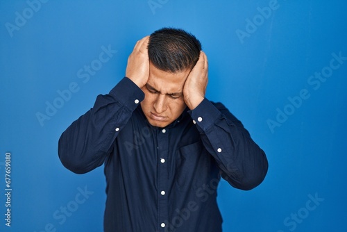 Hispanic young man standing over blue background suffering from headache desperate and stressed because pain and migraine. hands on head.