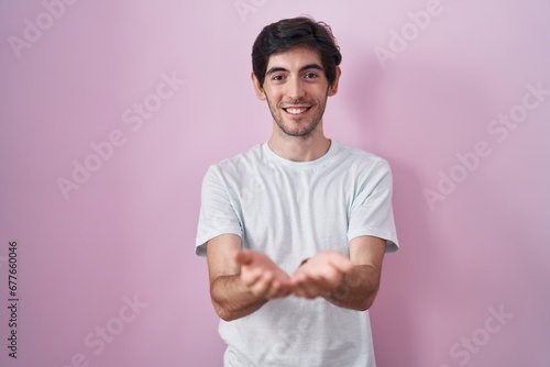 Young hispanic man standing over pink background smiling with hands palms together receiving or giving gesture. hold and protection