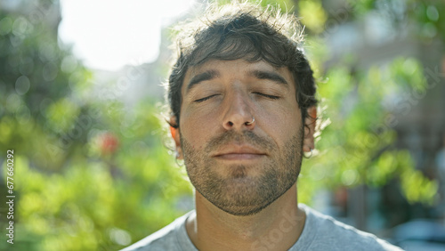 Hispanic young man with closed eyes, enjoying a breathing meditation in a sunny city park, a symbol of freedom and balance. blond beard, relaxed portrait, basking in the summer sunlight outdoors.