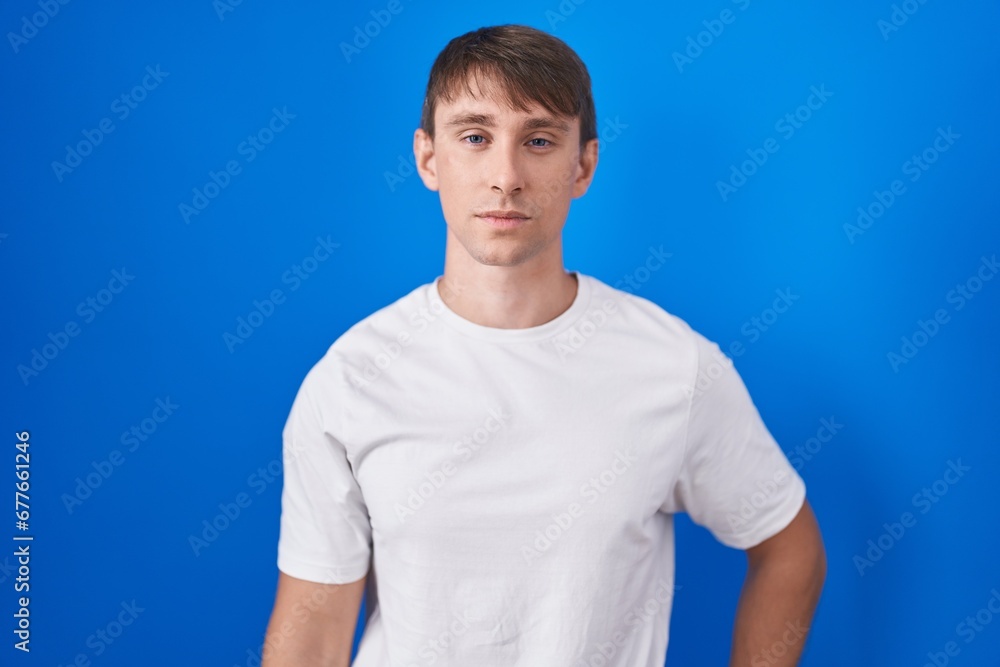Caucasian blond man standing over blue background relaxed with serious expression on face. simple and natural looking at the camera. Caucasian blond man standing over blue background relaxed with serious expression on face. simple and natural looking at the camera.