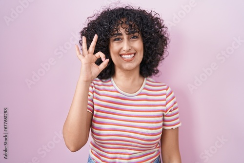 Young middle east woman standing over pink background smiling positive doing ok sign with hand and fingers. successful expression.