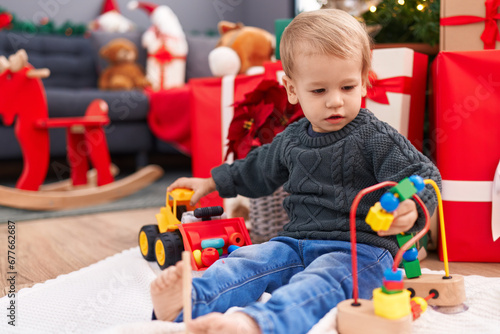 Adorable blond toddler playing with toys sitting on floor by christmas gifts at home