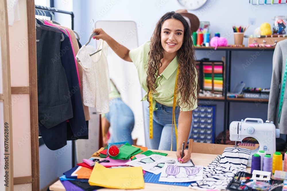 Fototapeta premium Young beautiful hispanic woman tailor smiling confident holding t shirt writing on notebook at clothing factory
