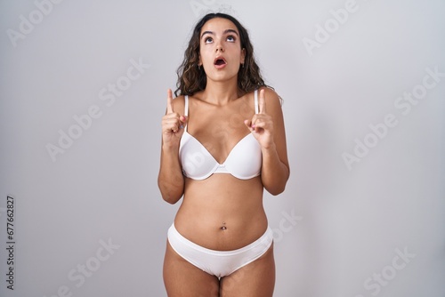 Young hispanic woman wearing white lingerie amazed and surprised looking up and pointing with fingers and raised arms.