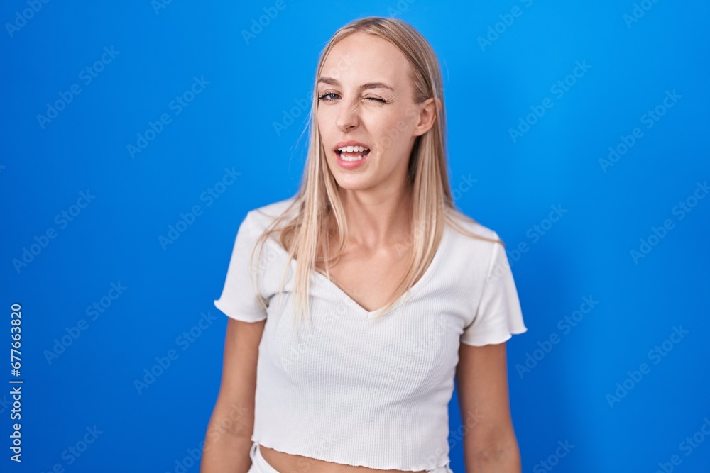 Young caucasian woman standing over blue background winking looking at the camera with sexy expression, cheerful and happy face. Young caucasian woman standing over blue background winking looking at the camera with sexy expression, cheerful and happy face.
