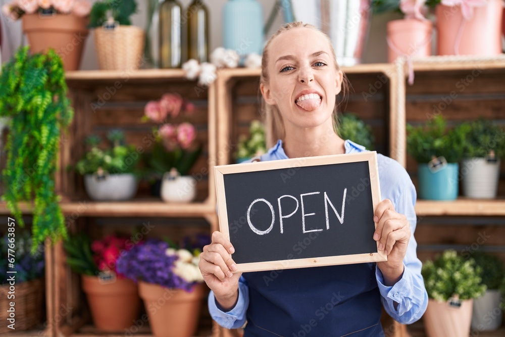 Young caucasian woman working at florist holding open sign sticking tongue out happy with funny expression. Young caucasian woman working at florist holding open sign sticking tongue out happy with funny expression.