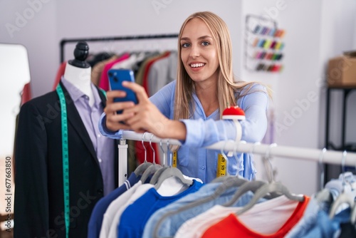 Young blonde woman tailor using smartphone leaning on rack at tailor shop