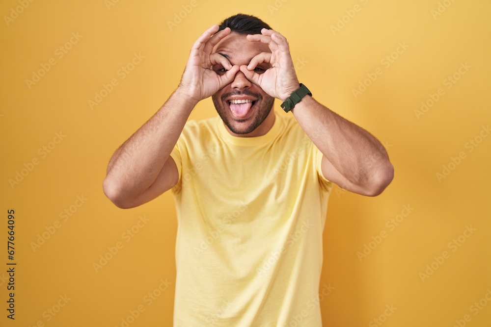 Young hispanic man standing over yellow background doing ok gesture like binoculars sticking tongue out, eyes looking through fingers. crazy expression. Young hispanic man standing over yellow background doing ok gesture like binoculars sticking tongue out, eyes looking through fingers. crazy expression.
