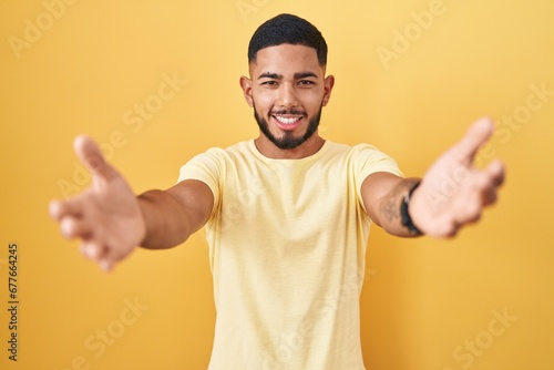 Young hispanic man standing over yellow background looking at the camera smiling with open arms for hug. cheerful expression embracing happiness. © Krakenimages.com