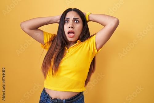 Young arab woman standing over yellow background crazy and scared with hands on head, afraid and surprised of shock with open mouth