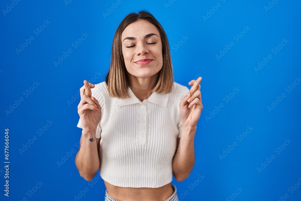 Hispanic woman standing over blue background gesturing finger crossed smiling with hope and eyes closed. luck and superstitious concept. Hispanic woman standing over blue background gesturing finger crossed smiling with hope and eyes closed. luck and superstitious concept.