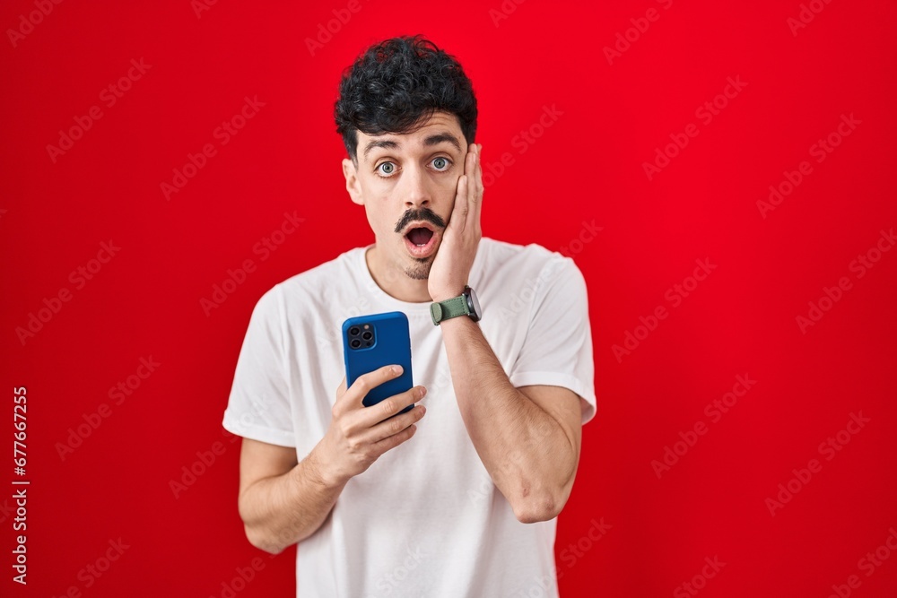 Hispanic man using smartphone over red background afraid and shocked, surprise and amazed expression with hands on face Hispanic man using smartphone over red background afraid and shocked, surprise and amazed expression with hands on face