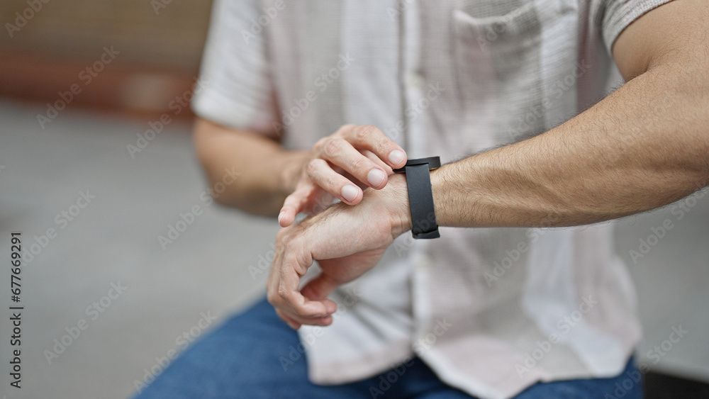 Young hispanic man touching watch sitting on bench waiting at street