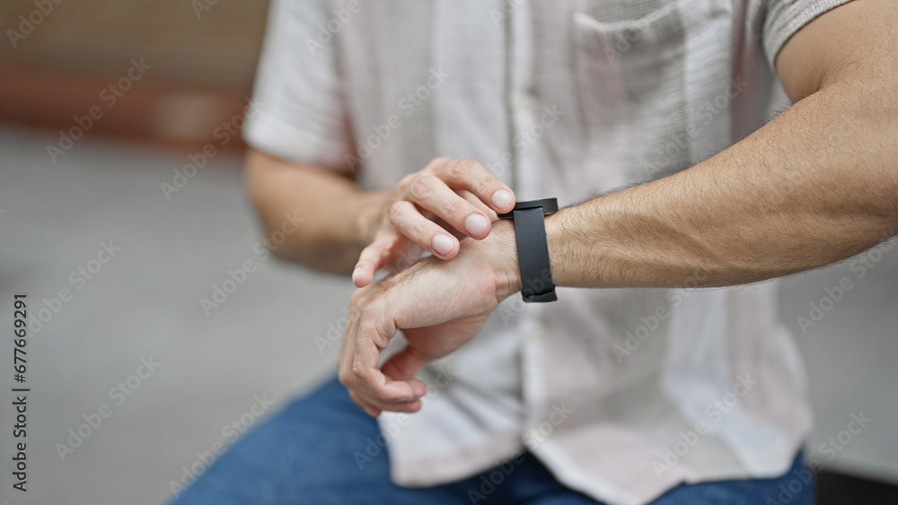 Young hispanic man touching watch sitting on bench waiting at street Young hispanic man touching watch sitting on bench waiting at street