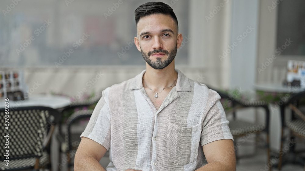 Young hispanic man sitting on bench with serious expression at coffee shop terrace Young hispanic man sitting on bench with serious expression at coffee shop terrace