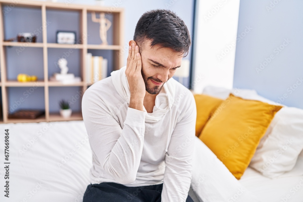 Young hispanic man suffering for ear pain sitting on bed at bedroom