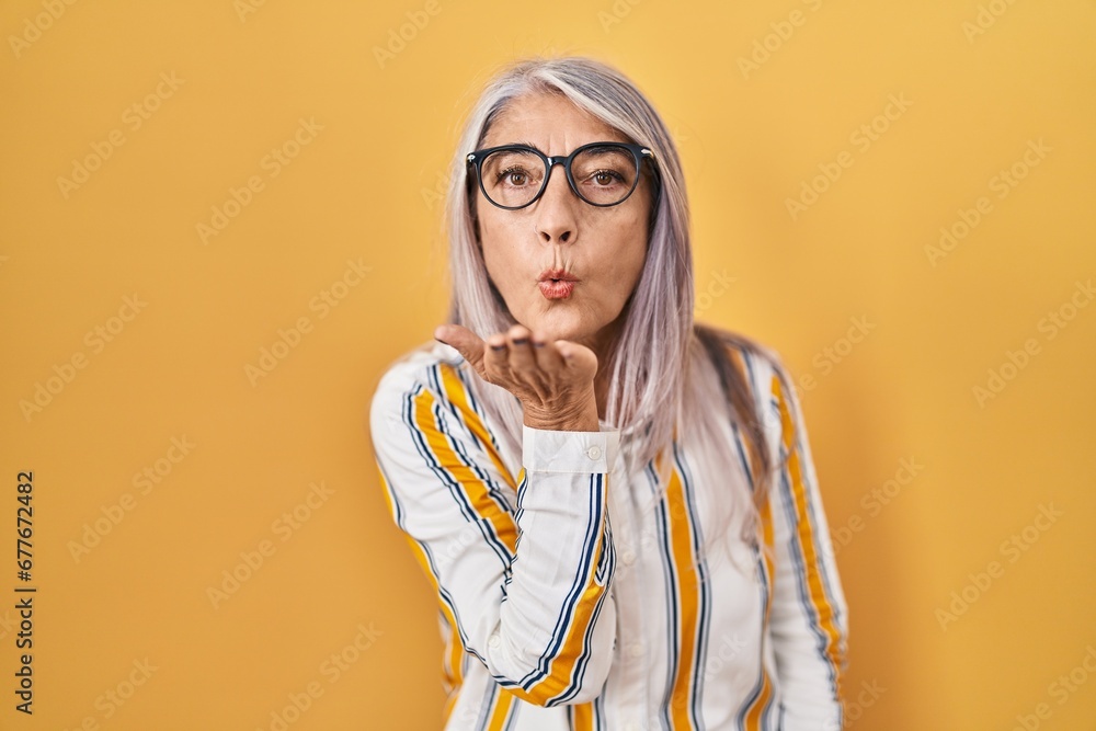 Middle age woman with grey hair standing over yellow background wearing glasses looking at the camera blowing a kiss with hand on air being lovely and sexy. love expression.
