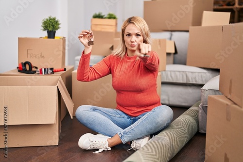Blonde woman holding keys of new home sitting on the floor pointing with finger to the camera and to you, confident gesture looking serious