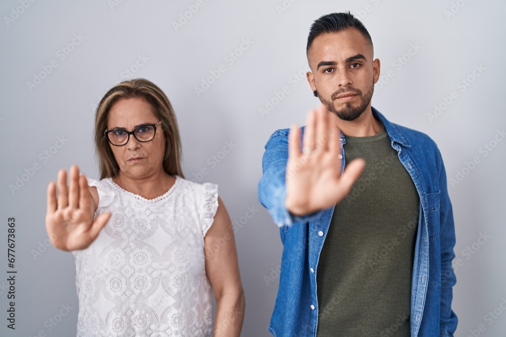 Hispanic mother and son standing together doing stop sing with palm of the hand. warning expression with negative and serious gesture on the face. Hispanic mother and son standing together doing stop sing with palm of the hand. warning expression with negative and serious gesture on the face.