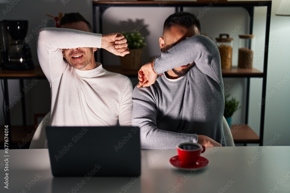 Homosexual couple using computer laptop smiling cheerful playing peek a boo with hands showing face. surprised and exited